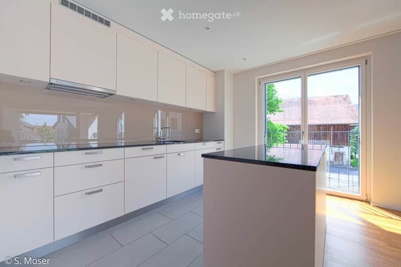 Modern kitchen with white cabinets, black countertops, and a central island. Features a large window with a view of the outdoor area.