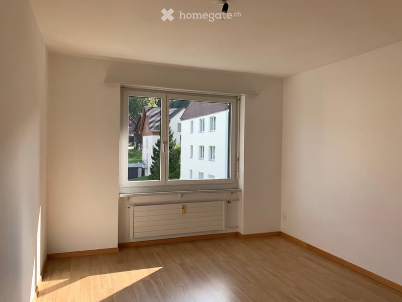 Empty room with wooden flooring, white walls, large window with view of neighboring buildings, radiator below window, and light fixture on ceiling.