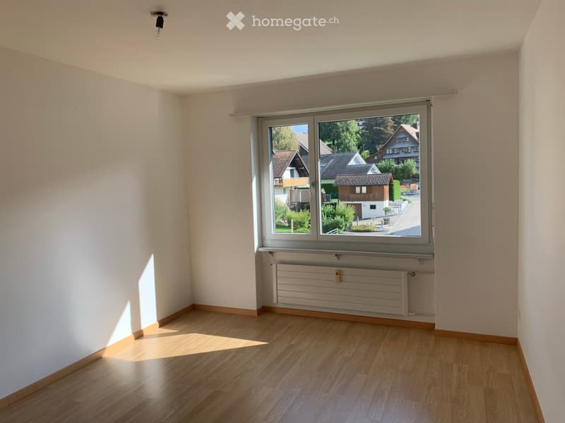 Empty room with wooden flooring, white walls, and a large window showing a view of houses and greenery outside.