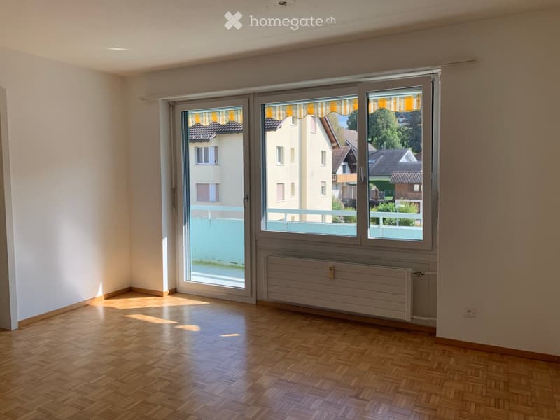 Empty room with wooden flooring, large windows with blinds, radiator, and a view of neighboring houses and a balcony.