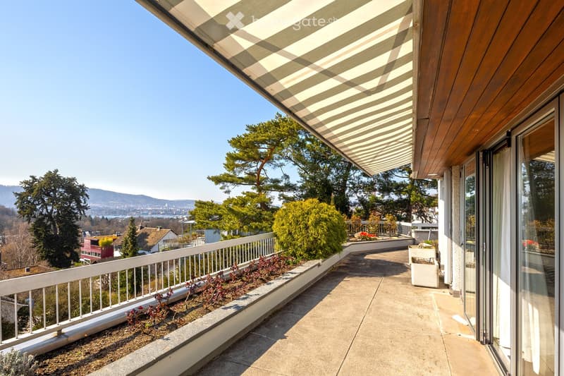 Balcony with white railing, striped awning, plants, and a view of the city and mountains