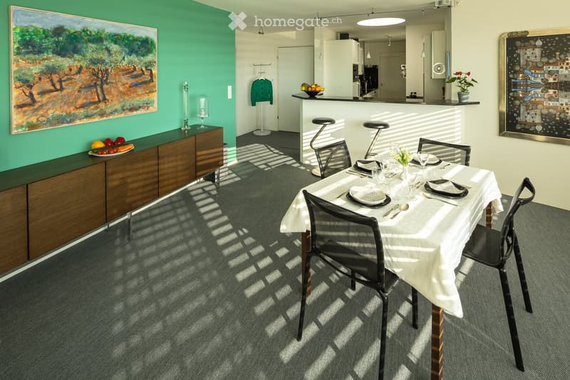 Dining room with a table, chairs, and a green wall with a painting. A counter with stools and a refrigerator is visible in the background.