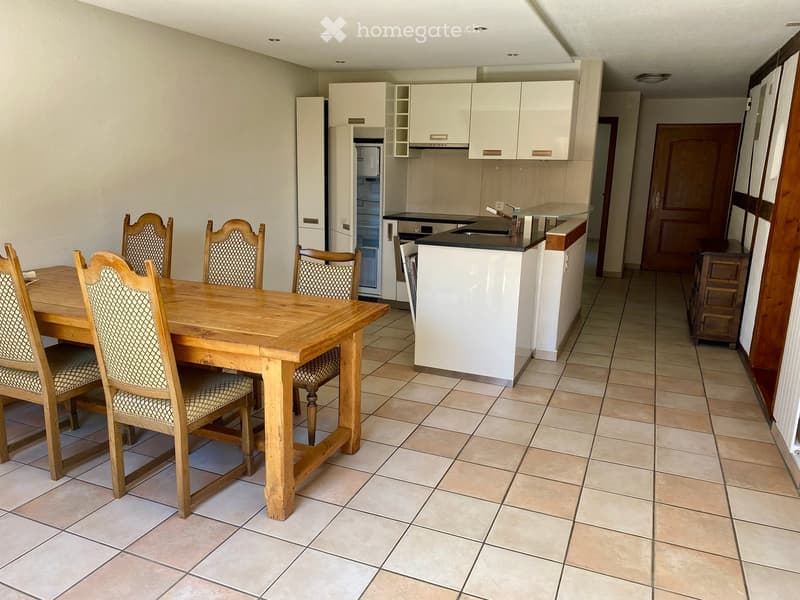 A well-lit dining area with a wooden dining table and six chairs, adjacent to a modern kitchen with white cabinets, a refrigerator, and a kitchen island with a sink.