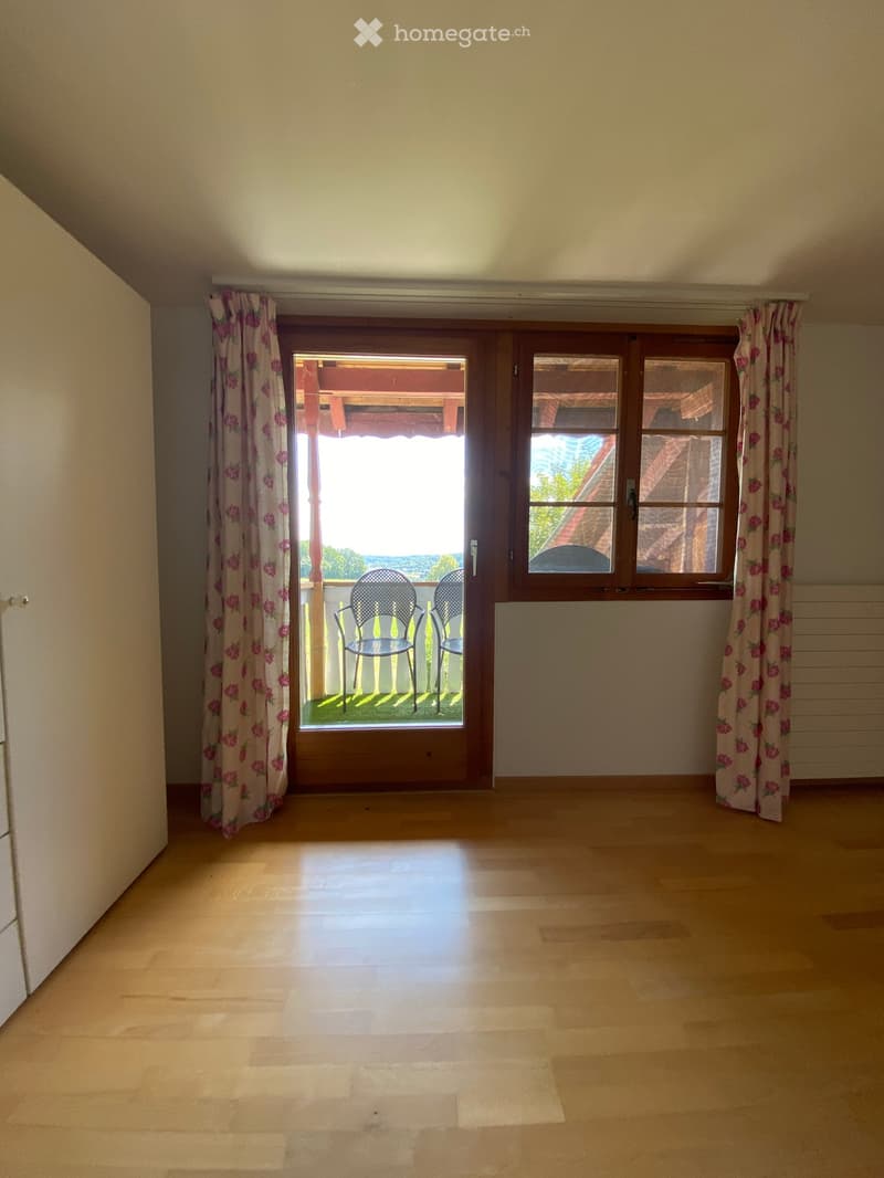 Empty room with wooden flooring, large window with floral curtains, and a door leading to a balcony with two chairs visible outside.