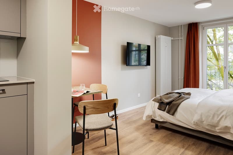 Modern, compact bedroom with a bed, a small table with chairs, a television mounted on the wall, and a curtained window. The room features a pendant light and a radiator.