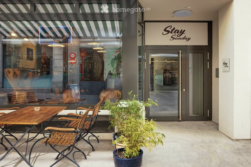 Outdoor seating area with wooden tables and chairs, a potted plant, and a storefront with a sign reading "Stay Sunday".