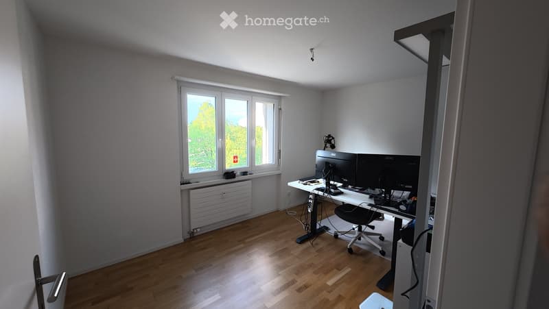 Bright, modern room with wooden flooring, large window, desk with dual monitors, chair, and radiator below the window.