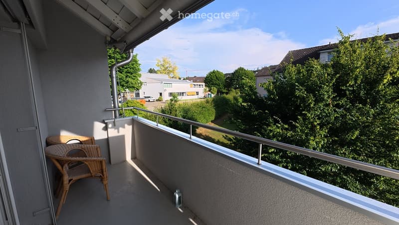 Balcony with a wicker chair and table, overlooking a green area with trees and a building in the background.