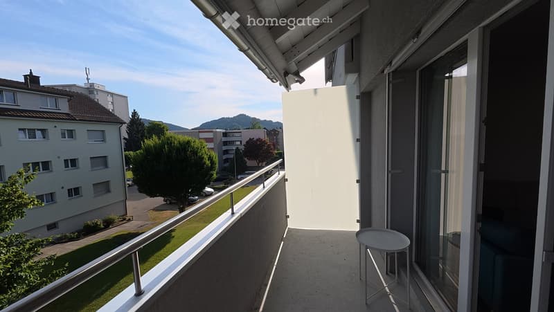 Balcony with metal railing, small white stool, view of street and buildings.