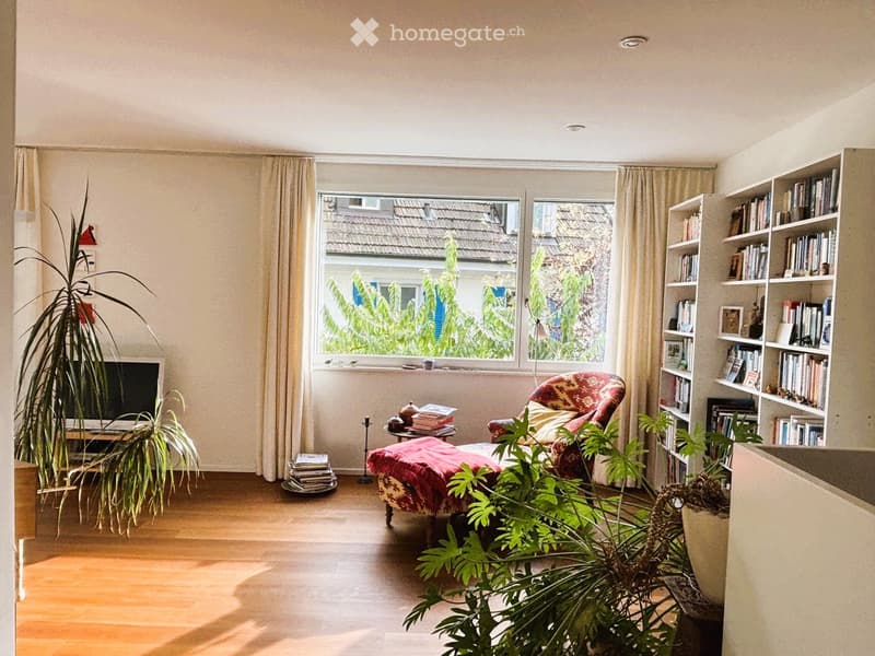Bright living room with wooden flooring, large window with view, sofa, bookshelf, television, and plants.