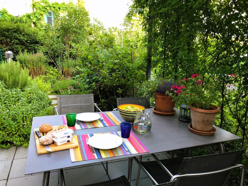 Outdoor dining setup with a table, chairs, plates, cups, and potted plants in a lush garden setting.