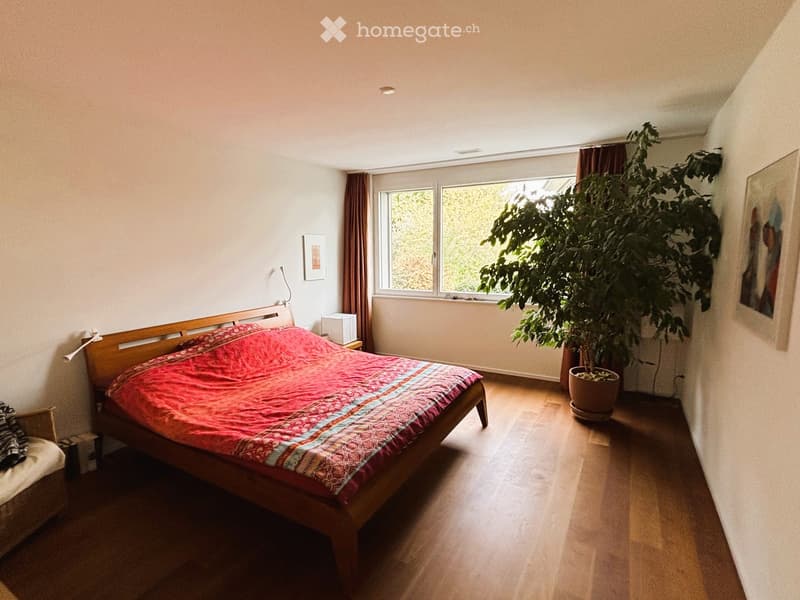Bright bedroom with wooden floor, large window with curtains, bed with red and blue patterned bedspread, nightstand, chair, potted plant, and picture frame on wall.