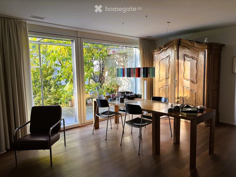 Bright dining area with wooden table, chairs, armchair, large windows, and a tall wooden cabinet.