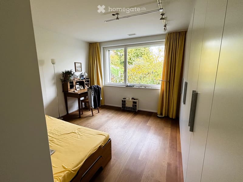 Bright bedroom with wooden flooring, large window with yellow curtains, bed with yellow bedding, desk with chair, small wine rack, and decorative items.