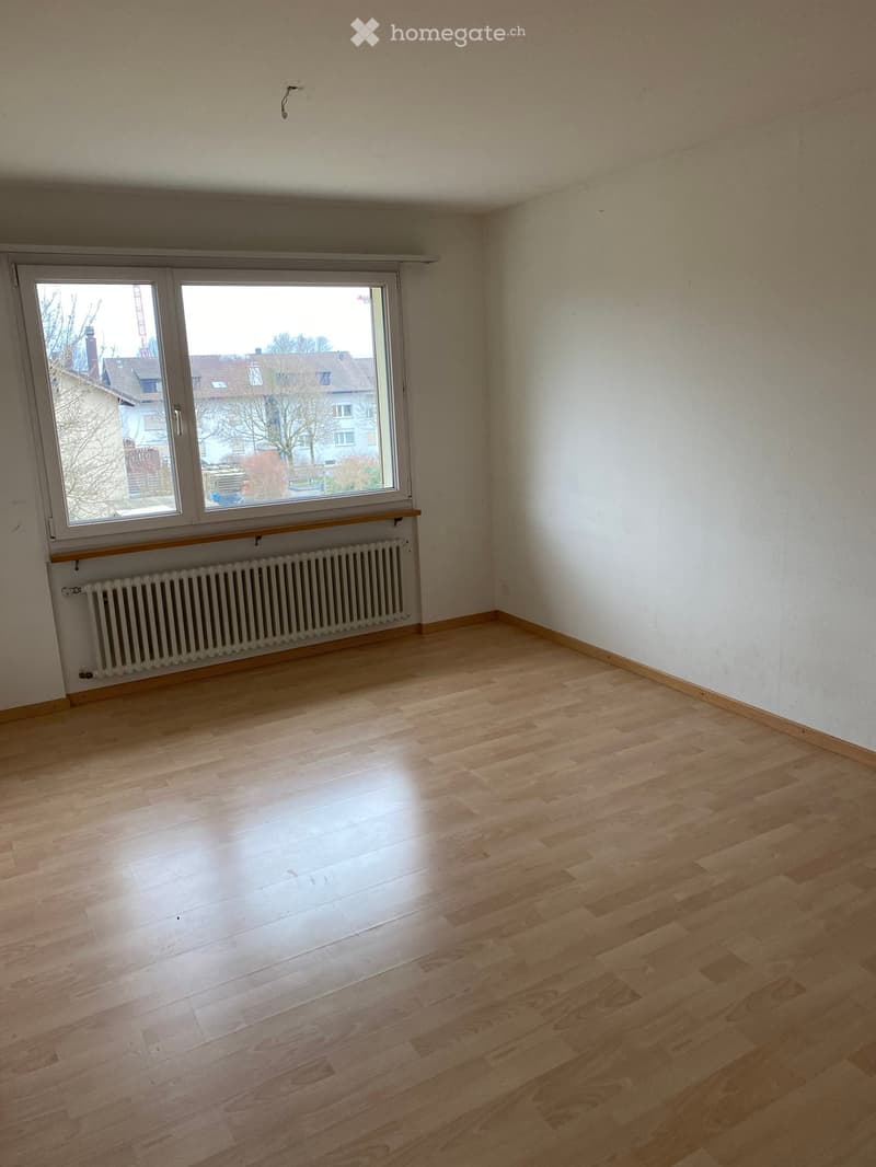 Empty room with wooden flooring, white walls, large window with radiator below, and visible outdoor view.