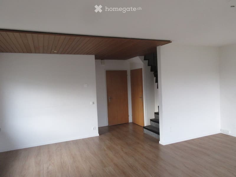 Empty room with wooden flooring, white walls, and a staircase leading upwards. Two wooden doors are visible in the background.