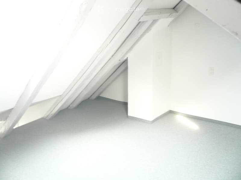 Empty attic space with exposed wooden beams and a sloped ceiling, featuring a smooth grey floor and white walls.