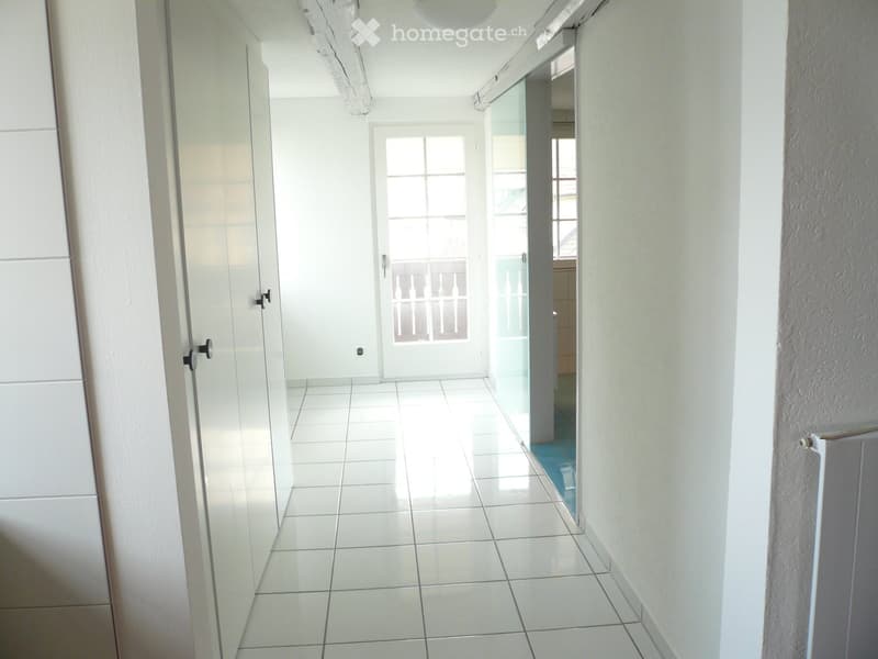 A clean, white-tiled hallway with a glass door leading to a balcony area. The walls are painted white, and the floor is covered with large, square tiles.