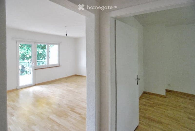 Empty room with wooden floor, white walls, open door to another room, sliding glass door leading to a balcony