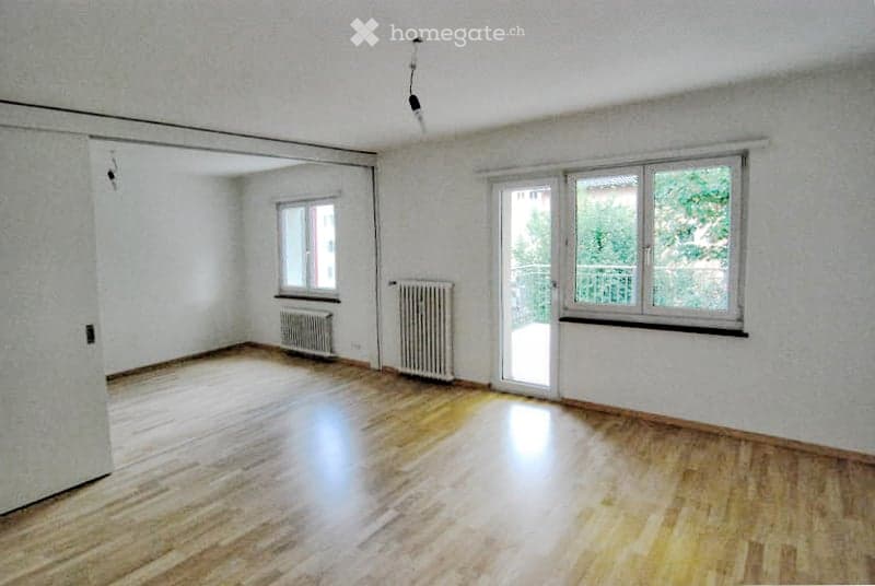Empty room with wooden flooring, white walls, and large windows. Radiators are visible along the walls, and there is a sliding glass door leading to a balcony.