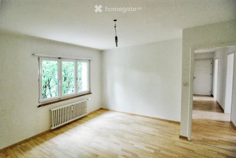 Empty room with wooden floor, white walls, large window, radiator