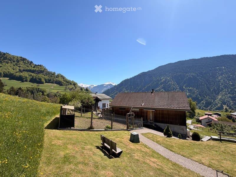 A wooden house with a tiled roof, surrounded by a grassy lawn and a mountainous landscape in the background.