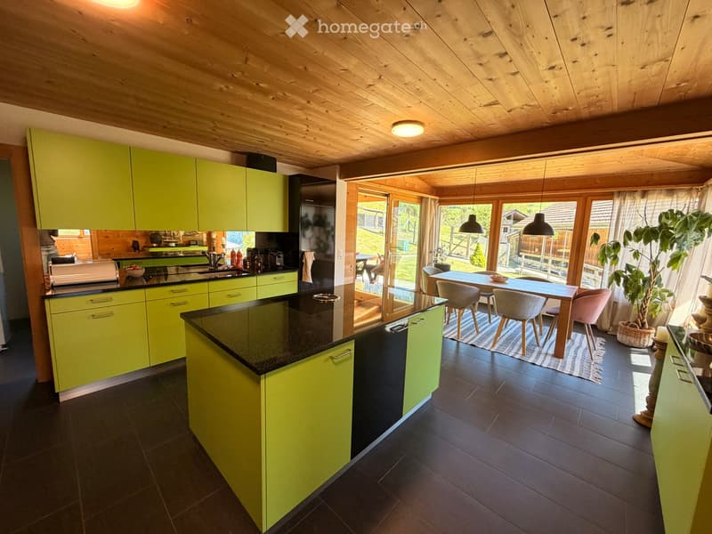 Bright kitchen with green cabinets, black countertops, and a central island. The space features a sink, refrigerator, oven, microwave, coffee maker, and a dining area with a table and chairs. Large windows provide natural light and a view of the outdoor a