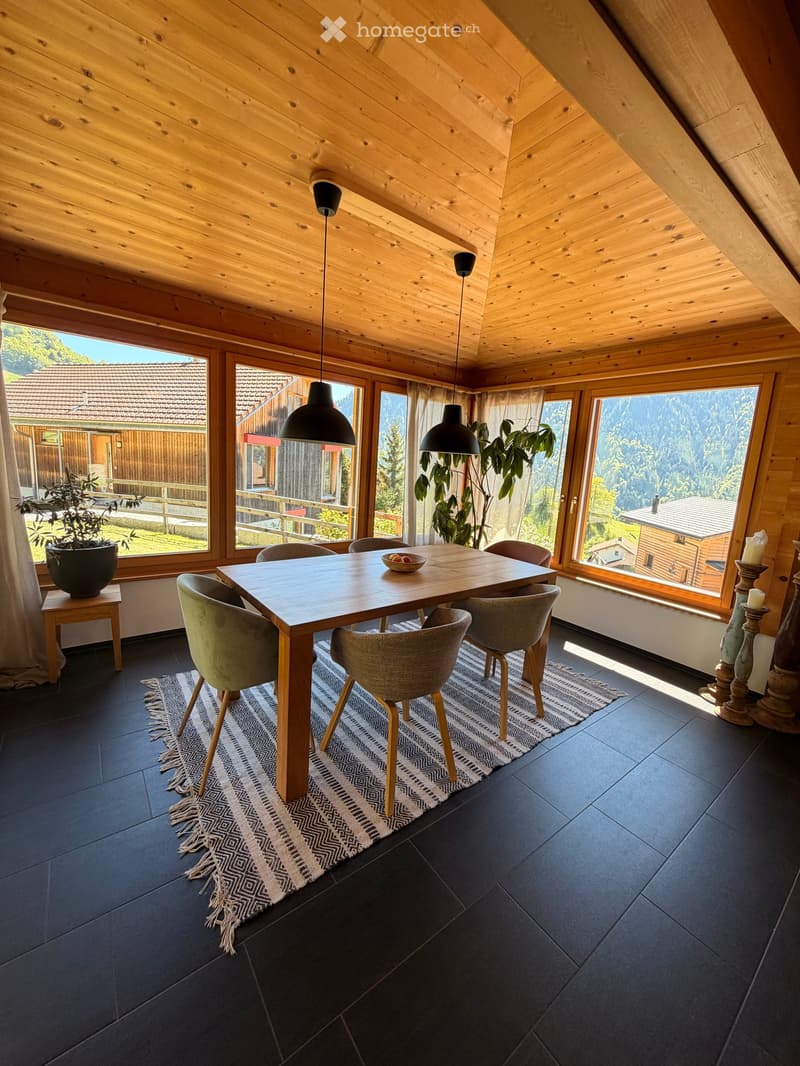 Bright dining area with wooden ceiling, large windows, dark tiled floor, and modern furniture including a dining table, chairs, pendant lights, and a patterned rug.
