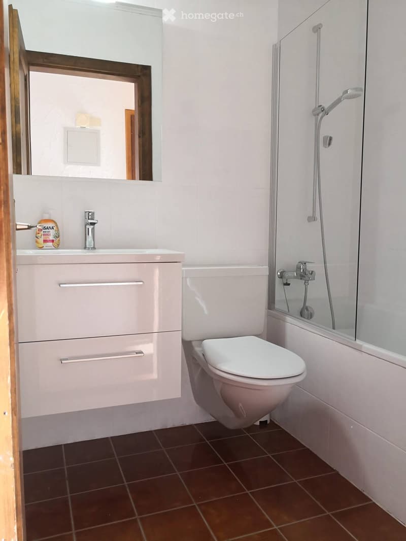 Modern bathroom with white tiles, a sink with a chrome faucet, a toilet, a shower with a glass enclosure, and a mirror above the sink. The floor is tiled in dark brown tiles.
