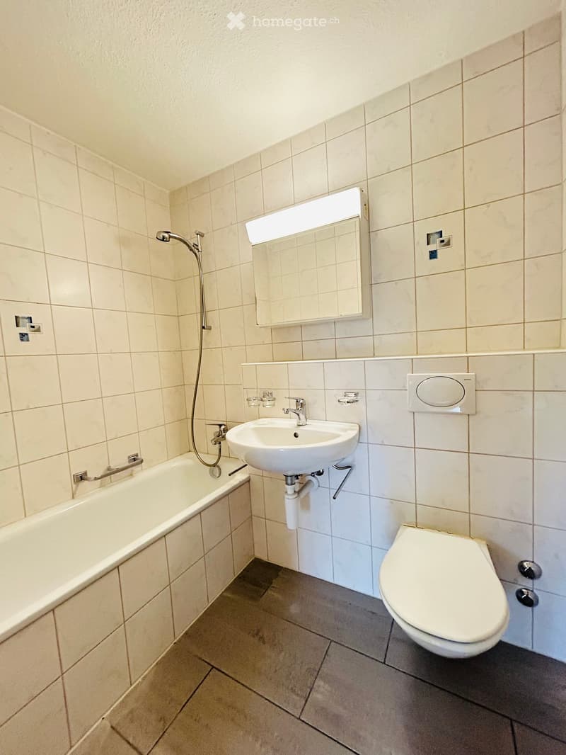 Modern bathroom with a bathtub, sink, toilet, and a large illuminated mirror above the sink. The walls are tiled in light beige, and the floor is tiled in dark gray.