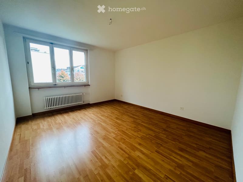 Empty room with wooden flooring, white walls, large window with view, radiator below window, and visible electrical outlet on wall.