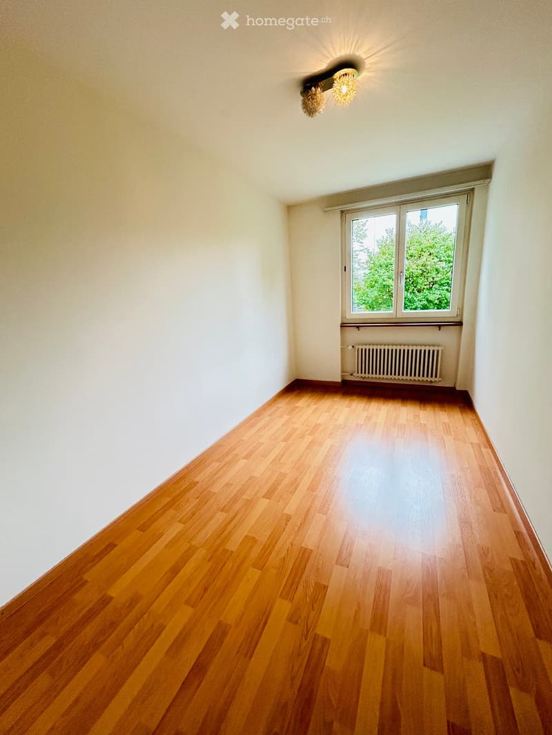 Empty room with wooden flooring, white walls, a window with white frame, and a radiator below the window.
