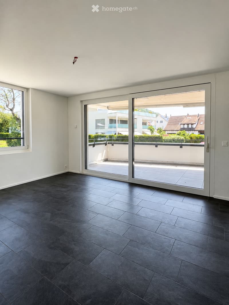 Empty room with dark grey tiled floor, white walls, large sliding glass doors leading to a balcony, and a window on the left side.