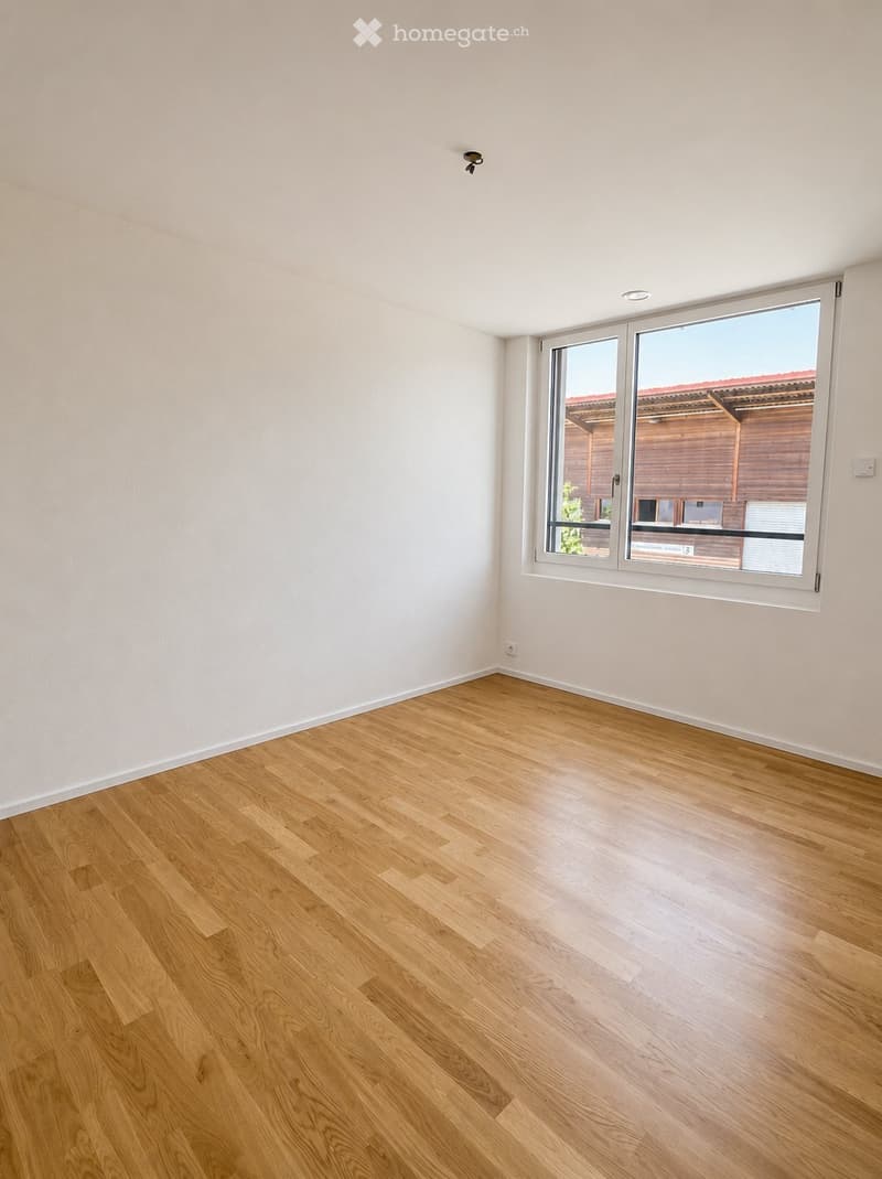Empty room with light wooden flooring and white walls, featuring a large window with a view of a wooden structure outside.