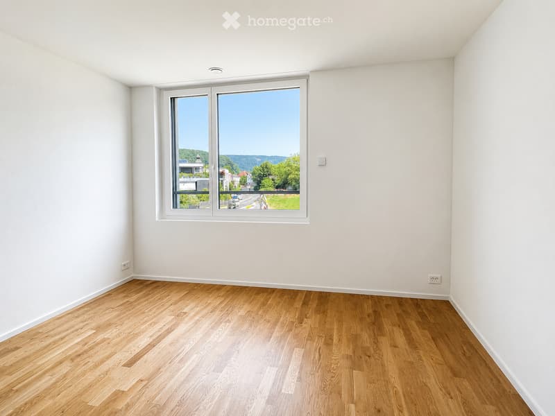 Empty room with light wooden flooring and white walls, featuring a large window with a scenic view of greenery and buildings.