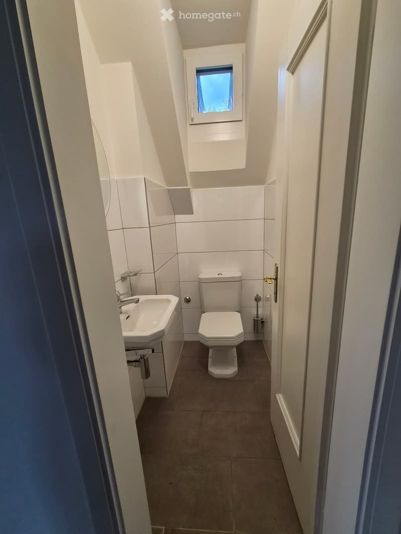 Modern, compact bathroom with white tiles, a sink, and a toilet. Features a small window and a door with a handle.