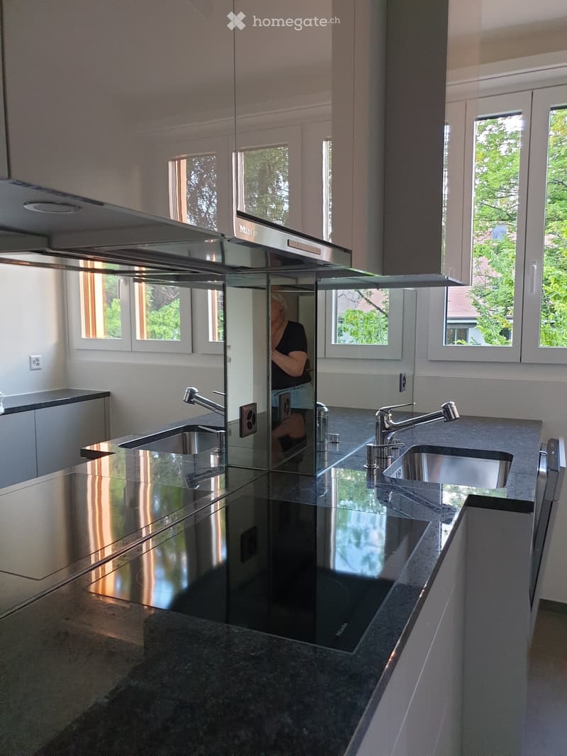 Modern kitchen with dark granite countertops, stainless steel sink, and sleek cabinetry. Large windows provide natural light.