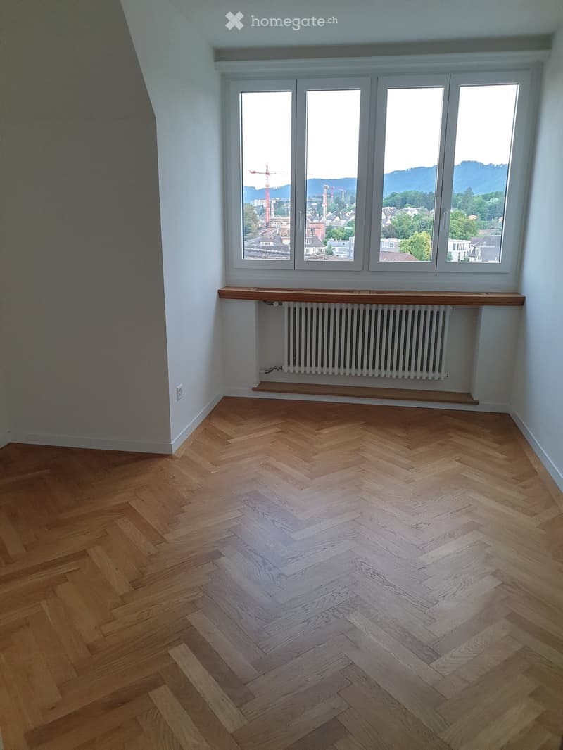 Empty room with herringbone-patterned wooden flooring, white walls, large windows offering a cityscape view, and a radiator with a wooden shelf above it.