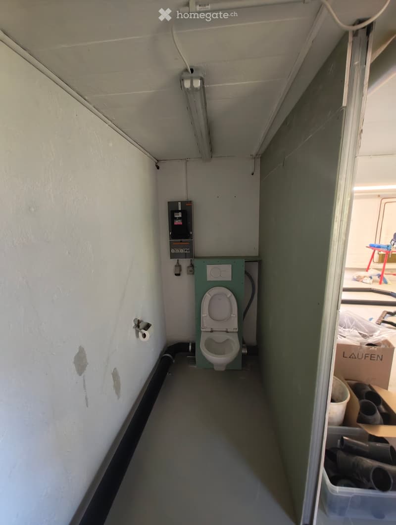 A small room containing a toilet, electrical box, pipework, and toilet roll. The room has white walls and a tiled floor.