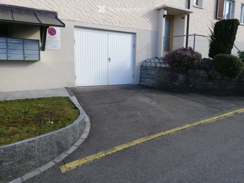 building entrance, white garage door, mailbox, yellow curb markings, grass, brick walls, plants