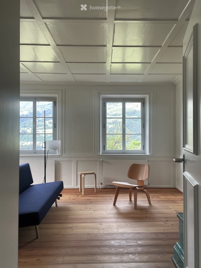 Bright, minimalist room with wooden flooring, large windows offering scenic views, a blue sofa, a wooden chair, a small stool, and a floor lamp.