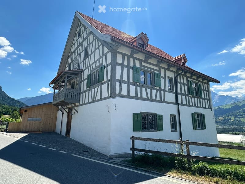 Traditional two-story house with white walls, dark wooden beams, green shutters, and a red-tiled roof, set in a scenic mountainous area.