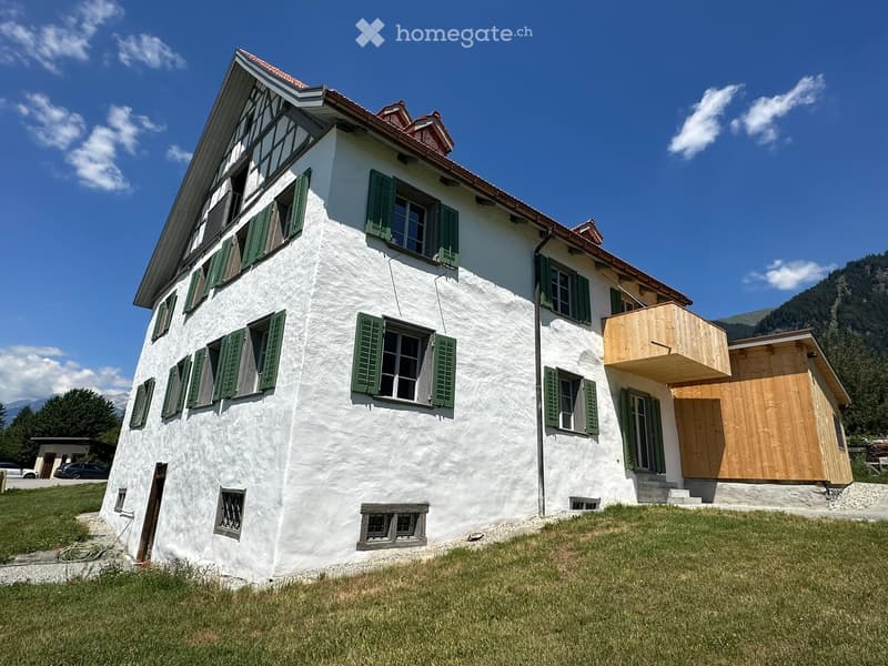 Two-story white house with green shutters, wooden balcony, and surrounding grassy area.