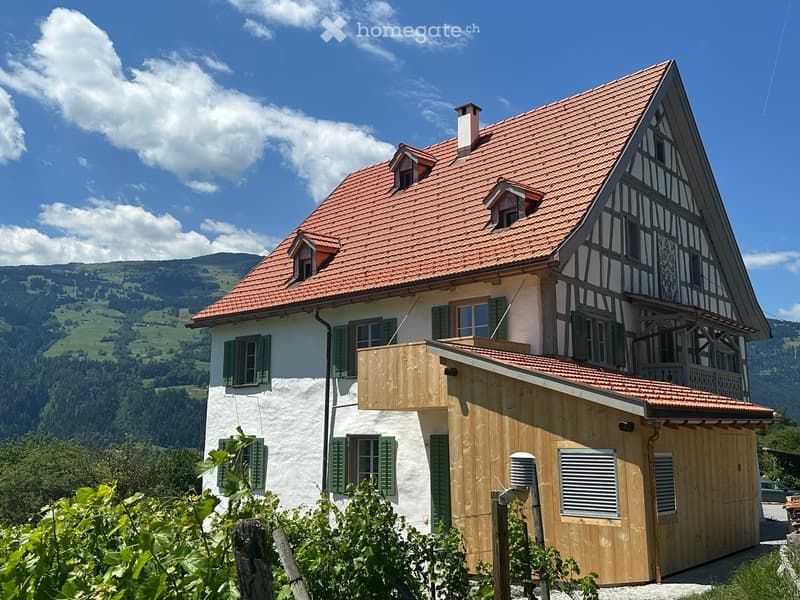 Traditional two-story house with red-tiled roof, white walls, green shutters, and wooden balcony overlooking a scenic valley.