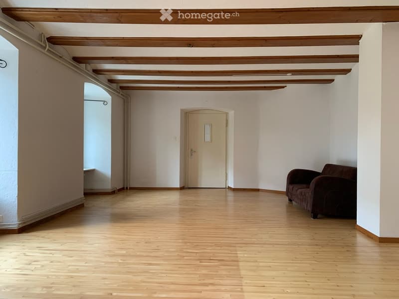 A spacious, well-lit room with wooden flooring and exposed wooden beams on the ceiling. The room features a single dark-colored sofa positioned against the wall, a closed door with a small window, and a visible hallway leading to another area.