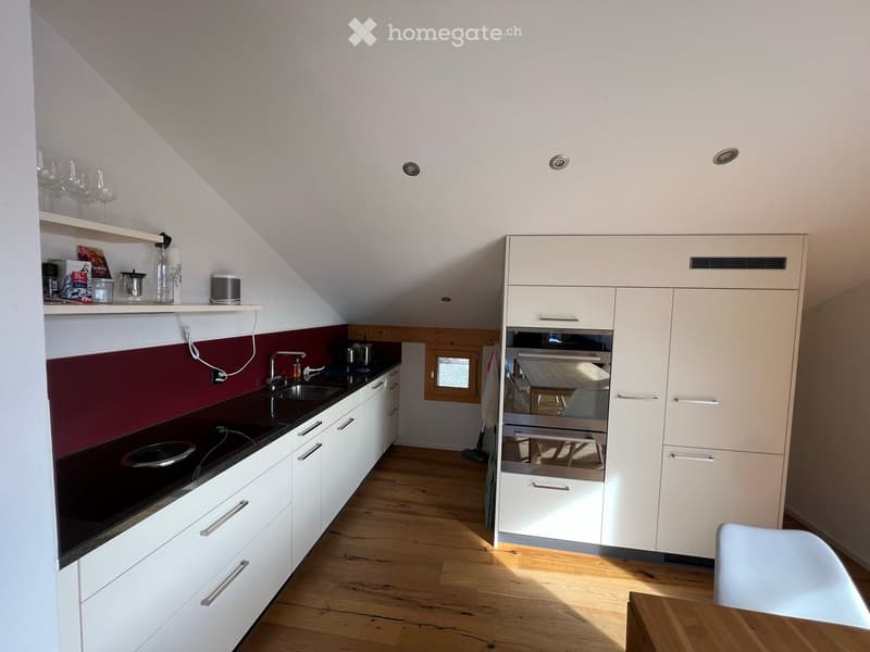 Modern kitchen with white cabinets, dark countertops, and wooden flooring. Features include a sink, faucet, built-in oven, and a small dining area with a table and chair.