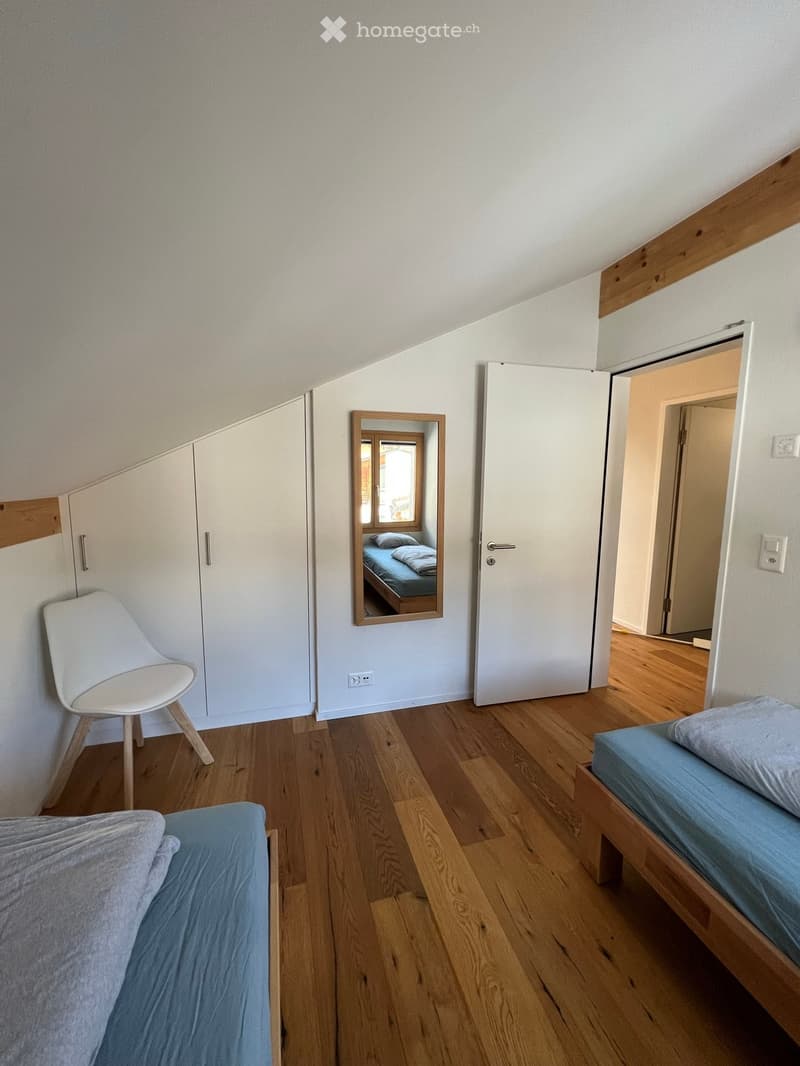Cozy attic bedroom with wooden floors, two single beds, a white chair, a mirror, and a built-in wardrobe.