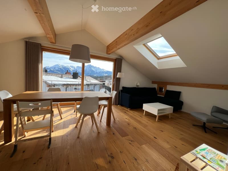 Bright, modern living room with wooden floors, large windows offering a mountain view, a sofa, coffee table, dining table with chairs, and a hanging lamp.