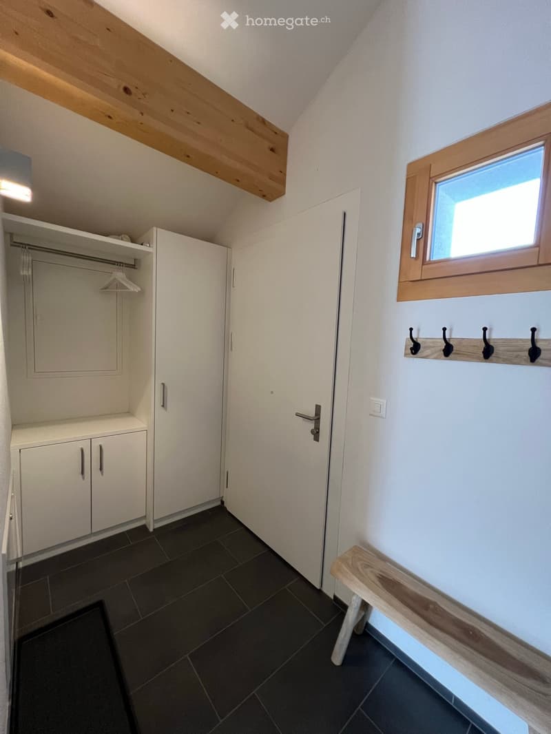 An empty room with white walls, dark tiled floor, built-in white closet with hanging space, small wooden bench, and a window with wooden frame.