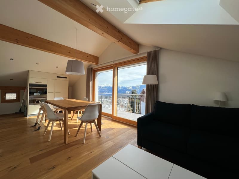 Bright, open living and dining area with wooden flooring, large windows offering a mountain view, a black sofa, a wooden dining table with white chairs, and a modern kitchen area with cabinets.